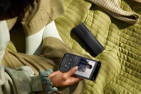A person sits on a green blanket holding their phone next to the black Roam 2 speaker.
