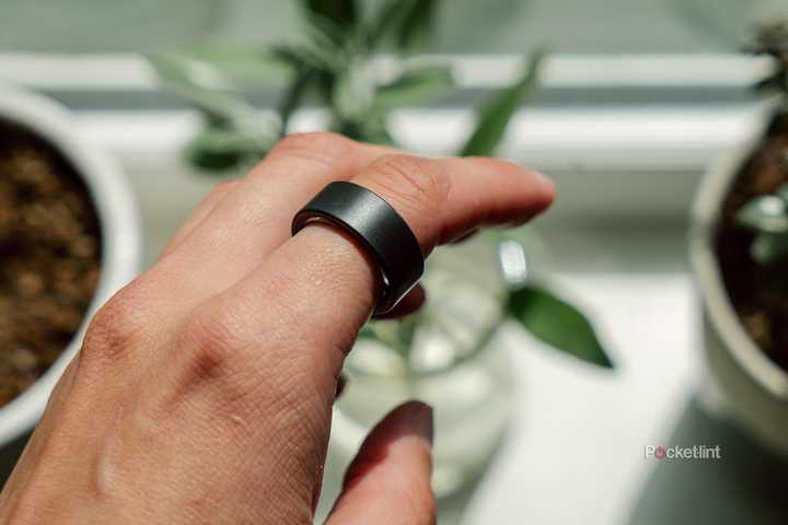 The Ultrahuman Ring Air on a finger held in front of a blurred out windowsill with plants. 